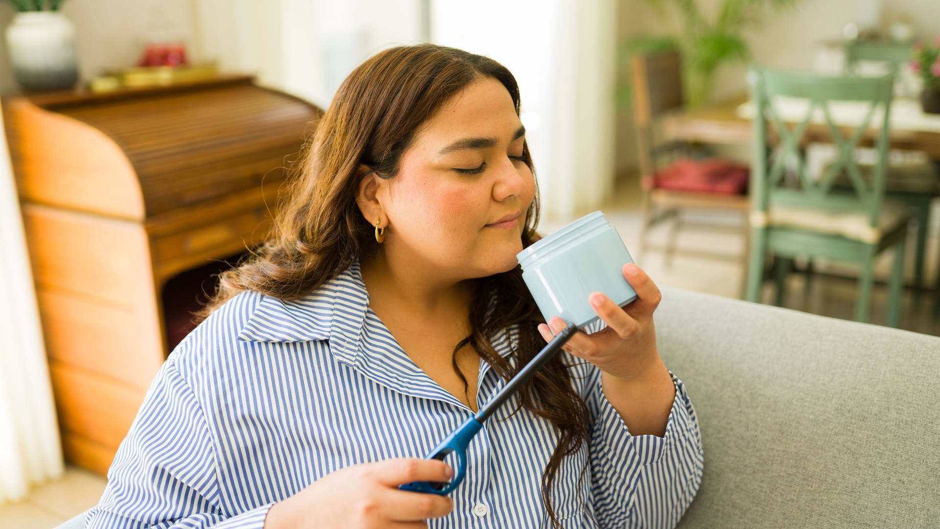 Woman enjoying self-care at home, sitting on a sofa, smelling a candle, embracing body positivity and comfort