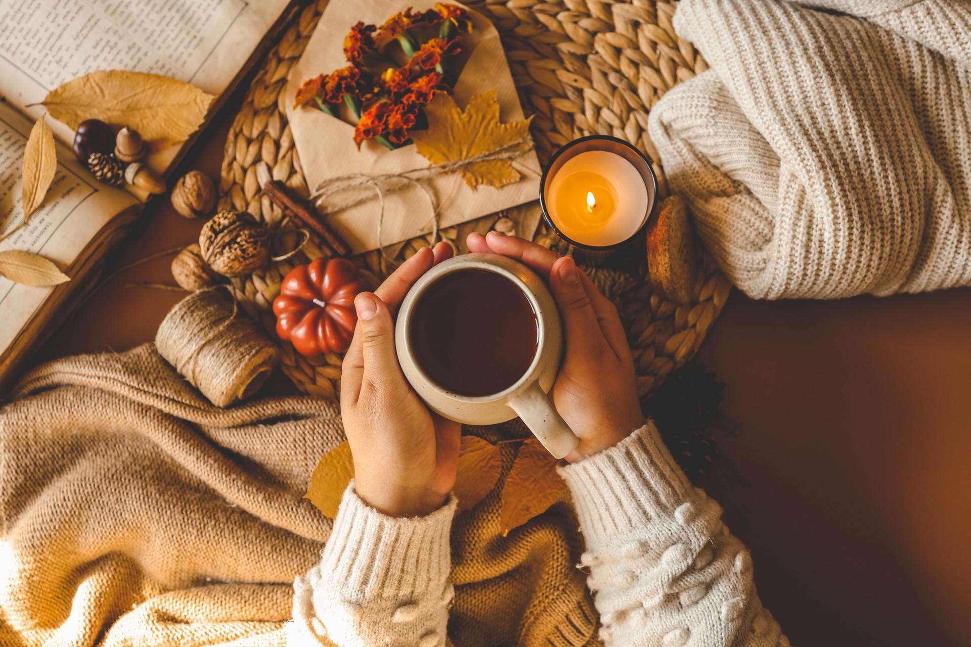 Cozy autumn flat lay featuring a warm drink held in hands, surrounded by fall elements such as a candle, pumpkin, twine, dried leaves, and a soft knitted sweater, evoking comfort and seasonal warmth