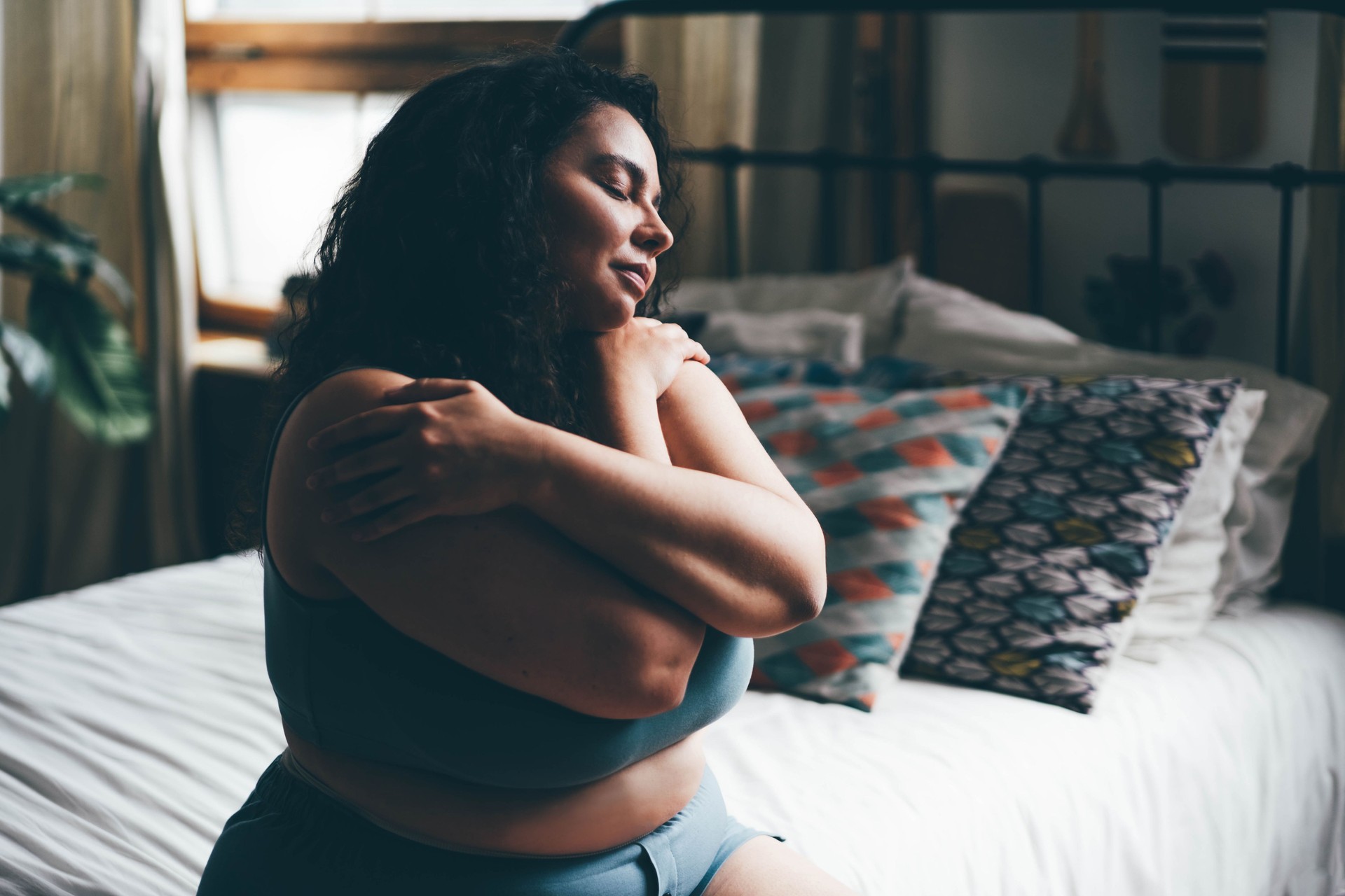 Curly haired overweight young woman in blue top and shorts with satisfaction on face accepts curvy body shape in stylish bedroom.