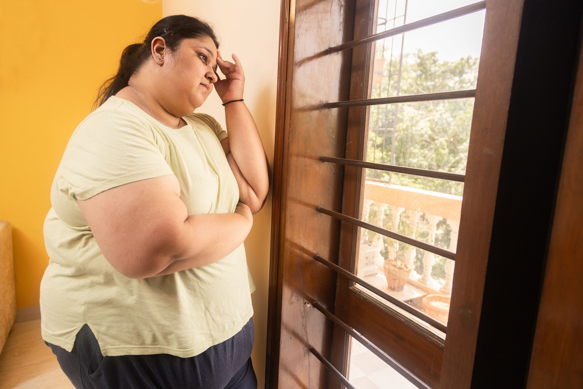 Stressed overweight fat indian woman thinking while standing near window at home. Plus size lady feels lonely and depression. Mental health problem due to obesity.